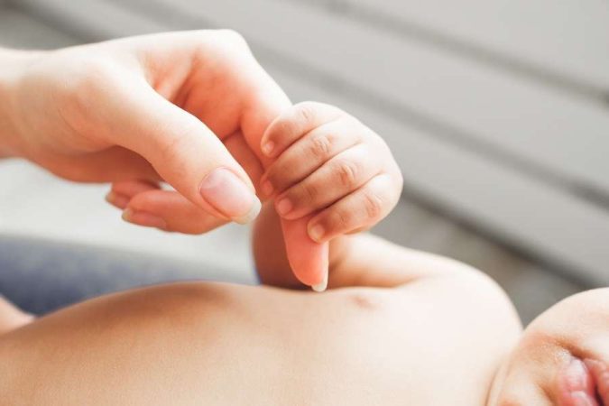 Une main adulte tient délicatement les petits doigts d'un bébé allongé sur le dos pour un moment de tendresse.