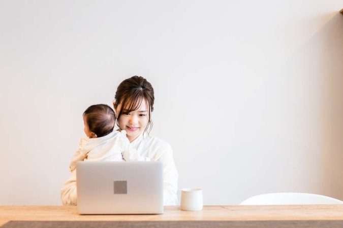 Une jeune mère sourit en travaillant sur son ordinateur portable tout en tenant son bébé, illustrant la réalité de la vie de famille et télétravail.
